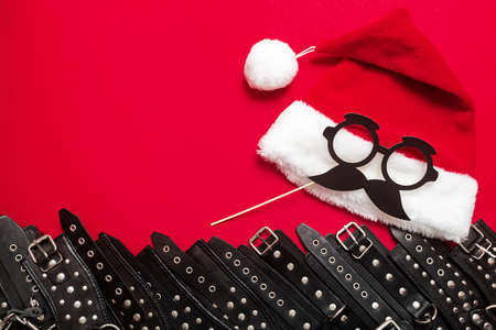Red color banner with black leather bracers and metal rivets for shackling hands or legs and performing sexual games. BDSM object and Santa hat with paper glasses. Background for adult products and sex toy store.の写真素材