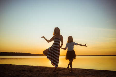 Mom with daughter in dresses are in a yoga pose on the lake at sunset. Leisure that strengthens the psychological health of the family and the body's immunity through hardening.の写真素材