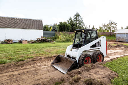 A skid steer loader clears the site for construction. Land work by the territory improvement. Machine for work in confined areas. Small tractor with a bucket for moving soil, turf and bulk materials.の写真素材