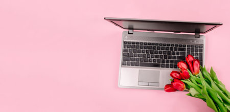 Bouquet of red tulips on pink paper background with a laptop on work place. Template for advertising or visualization of blog with copy space for text. Business card. Holiday certificate. Banner.の写真素材