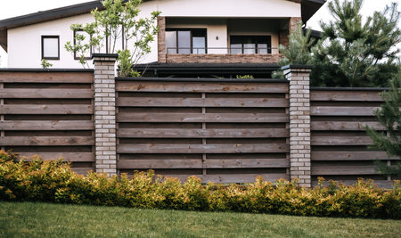 Section of a wooden fence with green plants. Evergreen hedge. Horizontal lines. Brick post. Scandinavian style of backyard. Western border. Home security barrier. Landscaping design. Unedged board.の写真素材