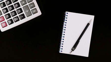 Large silver calculator with pen and white paper notebook on a black background. Copy space. Conceptual photo of calculations, accounting, computing, profit, loss, tax. Business card. Design mockup.の写真素材