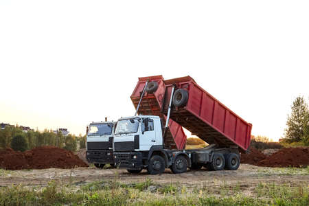 Two red dump trucks simultaneously lifted the bodies to unload the sand. Cargo transportation services. Large multi-ton truck. Unloading cargo. Construction site and machinery. Banner. Common view.の写真素材