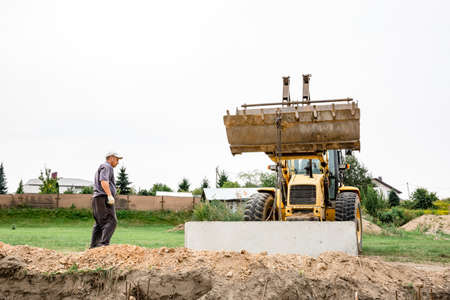 Wheel loader. Excavator install the foundation blocks into a trench. Retaining wall building. Construction site. Workers manage installing. Full HD footage video. Semkovo, Belarus - September 3, 2021.のeditorial素材