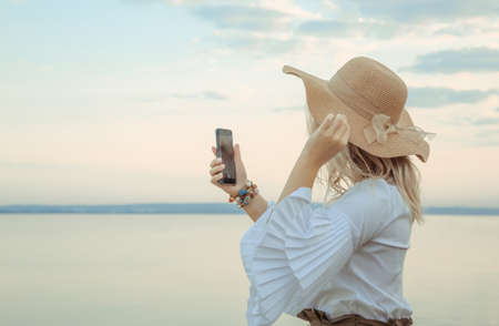 Portrait of a girl taking a selfie on the smartphone. Young beautiful 30 years woman caucasian appearance in straw hat with satin ribbon bow. Copy space. Summer lifestyle concept. Natural background.の写真素材