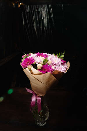 Large beautiful bouquet in the glass vase with chrysanthemums, gerberas and ferns in purple colors, packed in brown craft paper and isolated on a black background. Postcard for the holiday. Close-upの写真素材