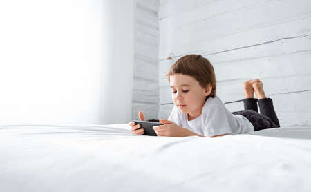 Little baby boy lays on his bed and plays with a smartphone. Close-up portrait. Holds in hands black cell phone. Concept of online education. White room and clothes. Telephone user. Save child visionの写真素材