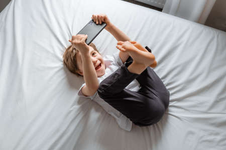 Little baby boy lays on his bed and plays with a smartphone. Close-up portrait. Holds in hands black cell phone. Concept of online education. White room and clothes. Telephone user. Save child visionの写真素材