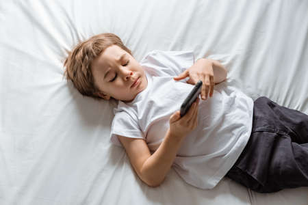 Little baby boy lays on his bed and plays with a smartphone. Close-up portrait. Holds in hands black cell phone. Concept of online education. White room and clothes. Telephone user. Save child visionの写真素材