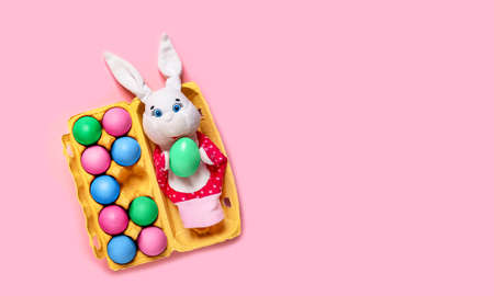Banner with Easter bunny and yellow packing with ten colorful eggs on pink background. Copy space, empty text place. Christian holiday card. Toy theater online course, hand puppet. Close-up mockupの写真素材