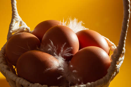 White handmade straw basket full of eggs and feather on yellow background. copyspace. Empty text...