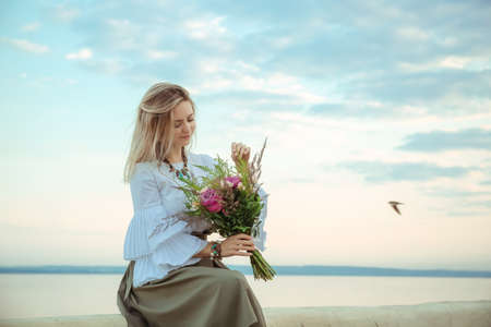 Portrait of young european woman sitting on the beach with flowers bouquet on the sea landscape background. Relaxing morning date walk. fashion. International woman or mother day. Harmony. happy mood.の写真素材
