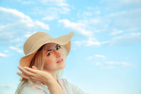 Portrait of young 30 year old european woman in straw hat and white dress looking at the camera on a cloudy sky background. beautiful girl. side view. profile. Romance mood. international woman day. hand.の写真素材