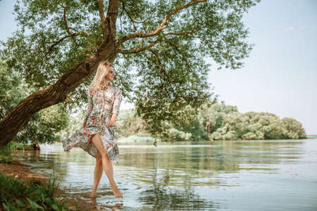 A young 30 year old woman in a long dress and barefoot walks along the beach lake. beautiful scenery. Feminine style of a European girl in sunglasses and straw hat. slim body. natural environment.の写真素材