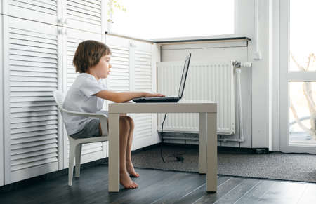 Boy sits at the table, uses laptop, typing on a keyboard, looks at the screen. Child does homework...