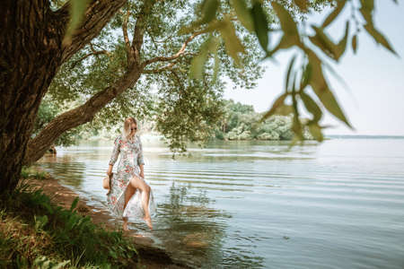 A young 30 year old woman in a long dress and barefoot walks along the beach lake. beautiful scenery. Feminine style of a European girl in sunglasses and straw hat. slim body. natural environment.の写真素材