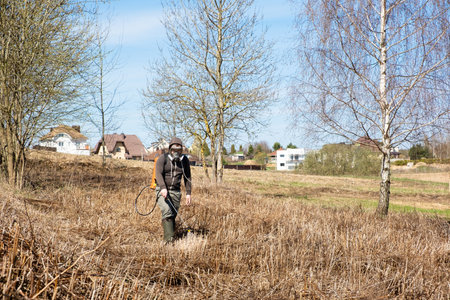 Farmer spraying pesticides and protects the field from weeds and ticks. Man with spray tank equipment. Tillage from ticks and insects. Springtime farm work. dry grass. water jet. manual labor. garden care.の写真素材
