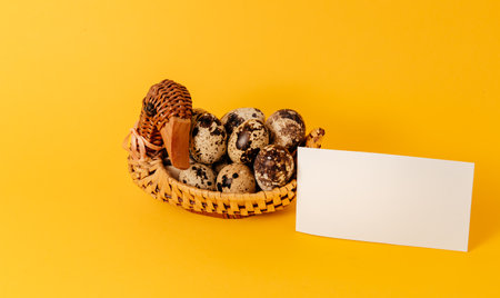Easter holiday mockup design. Handmade duck shaped straw basket with marble quail eggs on yellow background. interior decor. Healthy organic food. business card. paper note. Empty place for a message.の写真素材