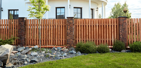 A new wooden fence with stone pillars finished with decorative stones. Landscaping. trimmed lawn and rock hill. young maple tree. natural materials. solid fence wall. House territory accomplishment.の写真素材