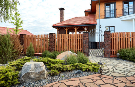 Entrance group of private house with wooden fence, stone pillars, lights, forged gate, paved path with red natural stone and flowerbed. Landscaping. boulders. Green planting territory accomplishment.の写真素材
