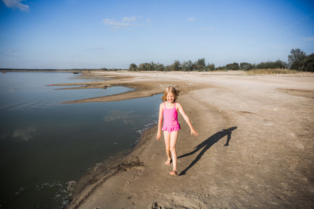 A little seven-year-old girl in a pink swimsuit runs along the lake in a good cheerful mood with a smile on her face. Summer lifestyle the holidays. Health tempering with water procedures and walks.の写真素材