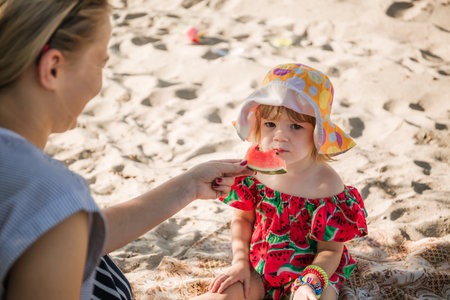 A beautiful young mother and little 2-year-old daughter are sitting on sand and eating a watermelon. Picnic on the beach. Sea travel holiday. A family care. Happiness lifestyle. love. close-up portrait.の写真素材