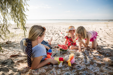 Mother and two daughters are sitting on sand eating the watermelon. Picnic on a beach. Sea travel holiday. Mommy care and love. Happy moments. Strengthening relationships parents and children. Morningの写真素材