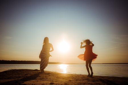 Silhouettes of mother and daughter in dresses jumping and dancing on the beach at sunset. Good relations between generations. Fun family pastime. Health promotion through outdoor activities. vacation.の写真素材