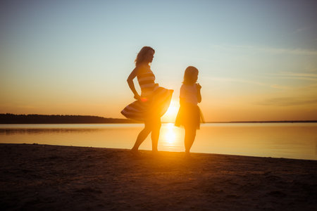 Silhouettes of mother and daughter in dresses jumping and dancing on the beach at sunset. Good relations between generations. Fun family pastime. Health promotion through outdoor activities. vacation.の写真素材