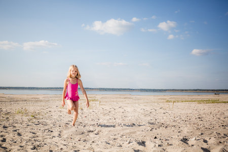 A little 7 year caucasian girl in a pink swimsuit running by sand on the lake in a good mood with a smile on face. Summer holiday lifestyle. Health tempering with swimming and walks. Weekend vibe.の写真素材