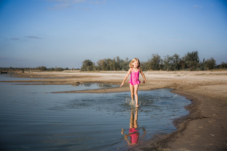 A little seven-year-old girl in a pink swimsuit runs along the lake in a good cheerful mood with a smile on her face. Summer lifestyle the holidays. Health tempering with water procedures and walks.の写真素材