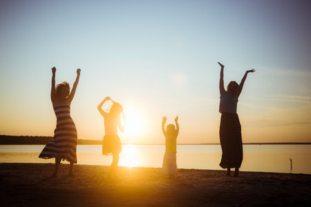 Silhouettes of children and mothers jumping and having fun on the beach in sunset light. Family weekend vibe. Good mood pastime among the generations. Beautiful natural landscape. Childhood happiness.の写真素材