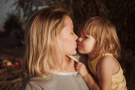 Mother holds 2 year daughter in her arms and kisses her in the rays of the sunset. Happy moments. Tactile contact. Family parent-child relationship. Love and happiness concept. Psychological comfort.の写真素材