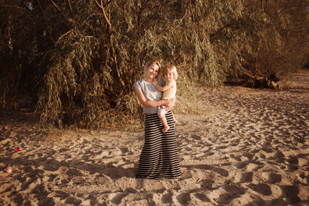 Mother holds 2 year daughter in her arms and kisses her in the rays of the sunset. Happy moments. Tactile contact. Family parent-child relationship. Love and happiness concept. Psychological comfort.の写真素材
