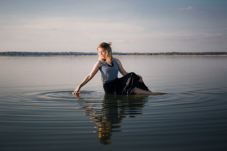 Portrait of beautiful young woman in dress sits in the water doing circles on water. Copy space. Fashion concept. Energy of water, earth and sun. Witch ritual. State of pleasure and bliss. Sexy pose.の写真素材