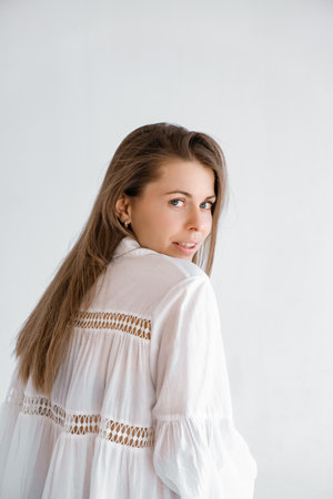 Beautiful young woman of Caucasian appearance looking over her shoulder. Smiling girl looks at the camera. White cotton dress with long sleeves in boho style. Oversized shirt. Femininity concept.の写真素材