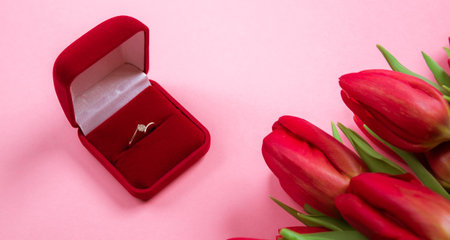 Diamond ring in box lais on a big bouquet of red tulips on pink background. Symbol of love. Jewelry...