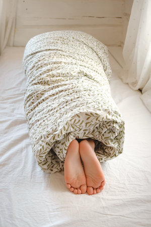 A child wrapped in a blanket with sticking out feet lies on the bed in the bedroom. Sunny morning...
