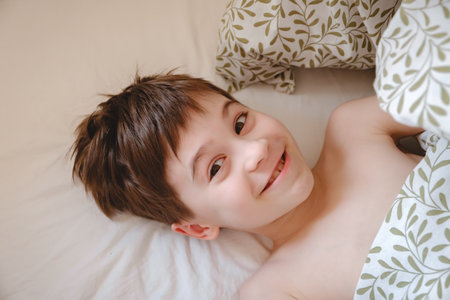 Smiling boy lying in bed looking at camera with mischievous look. Well-rested child in great mood view from above. Sleeping without pillow. Child sleeping without pajamas. Happyの写真素材