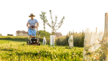 Person cutting grass in their yard with lawn mower. Mowing green grass. Summer gardening. Countryside lifestyle leisure. Spending free time at the garden. Sunny sunset. Slow motionの写真素材