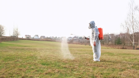 Man wearing protective suit treats the lawn from insect parasites using sprayer. Gardener in a full face mask respirator with personal protective equipment. Highの写真素材