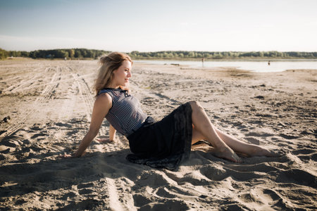 A young woman is sitting on sandy sea beach in a long black dress looking into the distance. Summer outdoor recreation. A meditative state. Sensual model enjoying the morningの写真素材