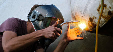 A skilled welder in protective mask welding the holes on a rusty metal tank. Sparks and glowing...