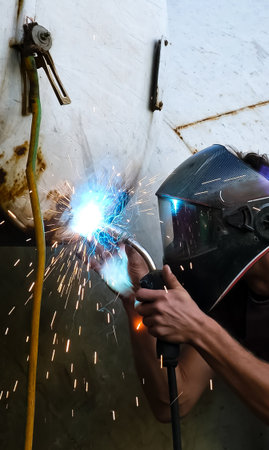A skilled welder in protective mask welding the holes on a rusty metal tank. Sparks and glowing...