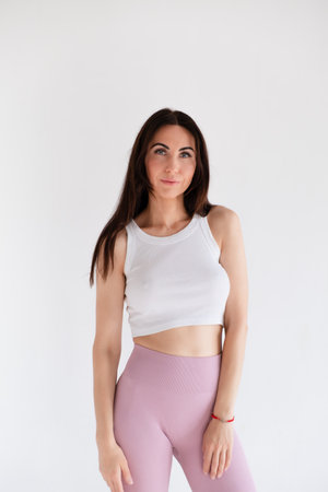 Young woman standing in activewear with abs and muscles, looking at camera isolated on white background. Caucasian 30s girl wears white top and pink leggings. Slim fit brunetteの写真素材