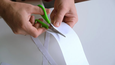 Male hands cutting a white sheet of paper into narrow strips with the green scissors on white background. Creating paper crafts. Art and labor lesson. Relieving stress and tensionの写真素材