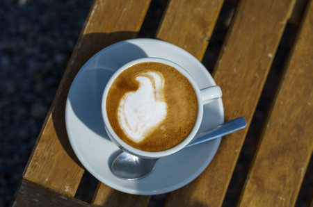 Hot cappuccino in a white mug with a spoon and saucer stands on a wooden table in the sunlightの写真素材