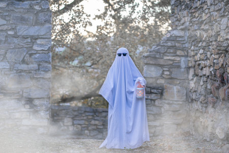 A child in a flowing white sheet walks along a stone wall wearing sunglasses and holding a glowing lantern in the middle of white smokeの写真素材