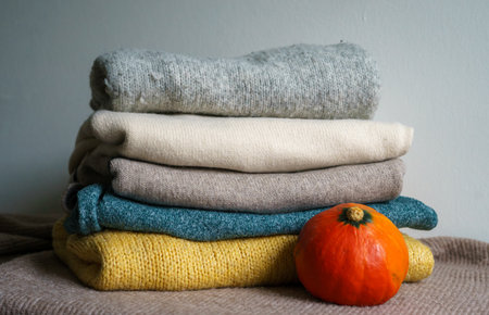 Stack of soft knitted sweaters in neutral tones with a small pumpkin, creating a warm and cozy autumn atmosphereの写真素材