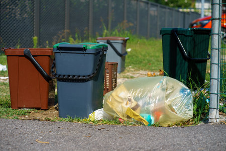 Household trash bins and a plastic garbage bag placed outdoors along a suburban street, illustrating waste management, recycling issues, and pollutionの写真素材